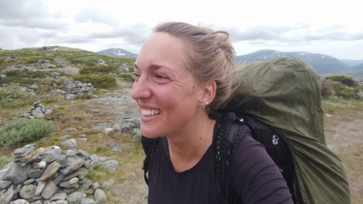 Es ist Johanna Geils mit einem Rucksack auf ihrer Reise zu sehen. Im Hintergrund erkennt man Berge und grüne Hügel.