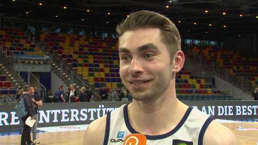 Der Basketballspieler Adrian Breitlauch steht während eines Interviews in einer Halle mit einem Mikrofon vor sich, die Tribünen im Hintergrund sind leer.