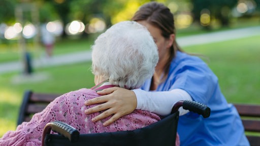 Eine Frau in blauer Pflegekleidung legt den Arm um eine Seniorin im Rollstuhl.