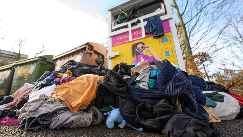 Gebrauchte Kleidungsstücke liegen vor einem Sammelcontainer für Altkleider