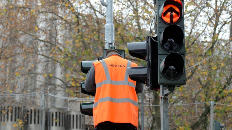 An einer Verkehrsampel führt ein Arbeiter mit Warnweste eine Reparatur aus.