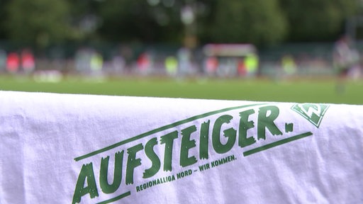 Im Vordergrund ist ein Shirt mit der Aufschrift "Aufsteiger" zu sehen, im Hintergrund Spieler auf einem Fußballfeld.