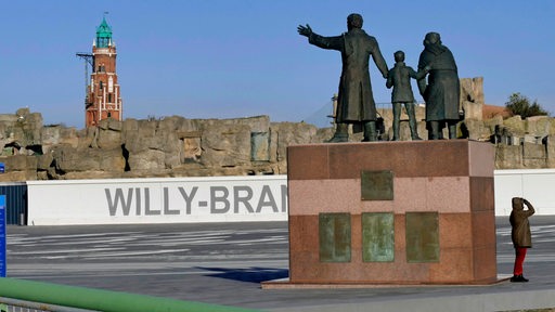 Eine Person steht neben einem Denkmal mit mehreren Personen auf einem Platz mit einem Leuchtturm.