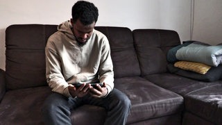 Der Mann Ayyub sitzt auf einem Sofa und schaut auf ein Handy.