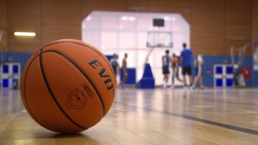 Ein Basketball liegt in einer Turnhalle, weiter weg im Hintergrund trainieren mehrere Spieler