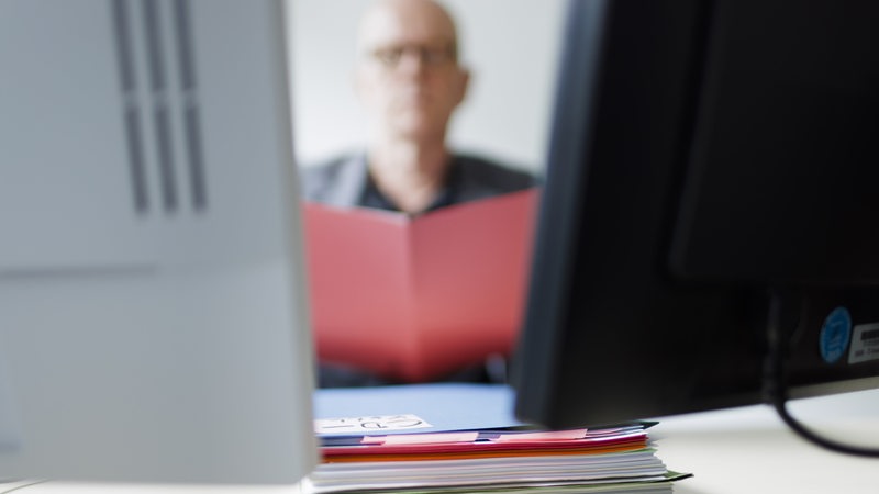 Unscharfe Person sitzt vor einem Aktenstapel und zwei Bildschirmen.