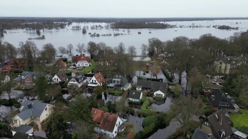 Ein überfluteter Straßenzug mit Wohnhäusern, im Hintergrund eine große Wasserfläche.