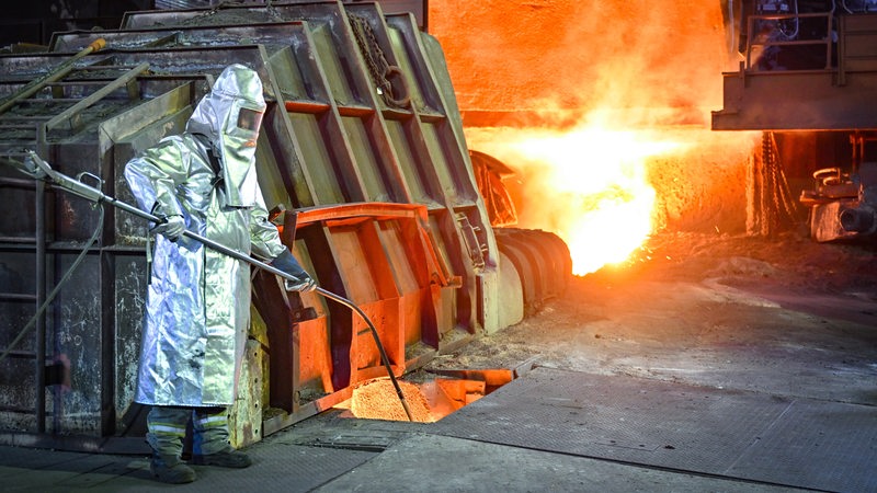 Ein Mitarbeiter in Schutzkleidung überwacht die Temperatur von Roheisen beim Abstich an einem Hochofen. 