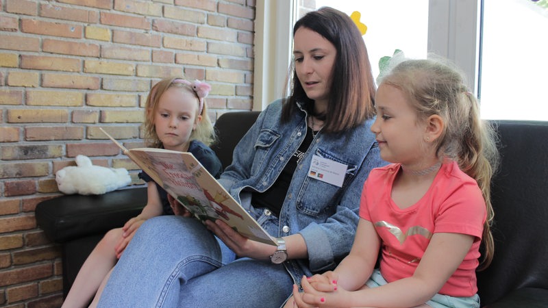 Eine Frau sitzt zwischen zwei Mädchen und liest aus einem Buch vor.