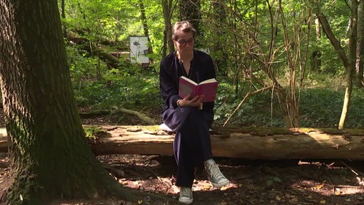 Buchautorin Katrin Seyfert sitzt auf einem Baumstamm im Wald und liest ihr Buch