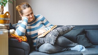 Eine Frau liest lächelnd auf dem Sofa ein Buch.