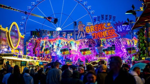 Das Fahrgeschäft Breakdancer ist auf dem Bremer Freimarkt hell beleuchtet. Es ist dunkel, Menschenmassen ziehen vor den Fahrgeschäften entlang.