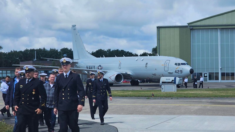 Mehrere uniformierte Personen laufen vor einem Flugzeug und einer Halle entlang.