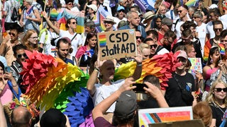 Viele Menschen sind zu sehen, einige tragen Kostüme in den Regenbogenfarben. Eine Person hält ein Schild mit der Aufschrift "CSD statt AfD".