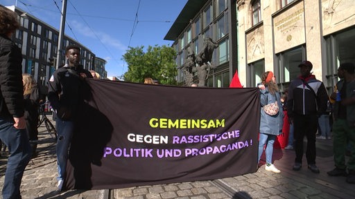 Zwei Personen halten ein Banner in den Händen, welches gegen rassistische Politik und Propaganda ist.