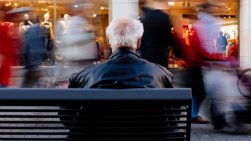 Ein Rentner sitzt auf einer Bank während vor ihm (mit Langzeitbelichtung) Menschen vorbeilaufen.