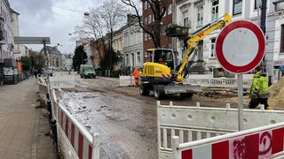 Eine Straße muss wegen eines Wasserrohrbruchs repariert werden. Im Vordergrund ist eine Baustellenabsperrung zu sehen und im Hintergrund arbeitet ein Bagger mit einem Bauarbeiter.