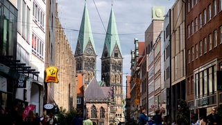 Zahlreiche Menschen sind bei sonnigem Wetter auf der Obernstraße in der Innenstadt unterwegs, während im Hintergrund der historische Bremer Dom zu erkennen ist. 