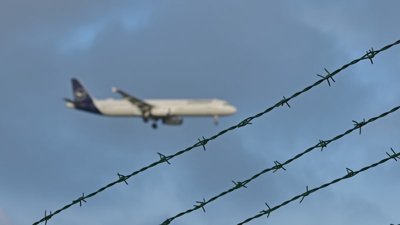 Ein Flugzeug ist am Himmel zu sehen. Im Vordergrund ist Stacheldraht zu sehen.