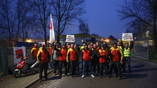 Briefträger und Briefträgerinnen stehen vor einer Halle und haben Streik-Plakate in der hand