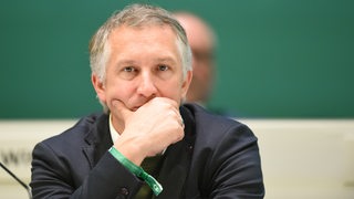 Werders Geschäftsführer Sport Frank Baumann sitzt nachdenklich bei der Mitgliederversammlung auf dem Podium und stützt nachdenklich den Kopf auf die Hand.