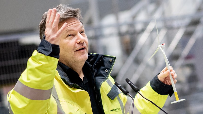 Ein Mann in gelber Jacke hält ein Windrad-Modell in der Hand.