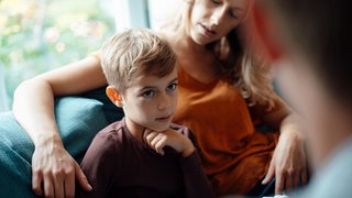 Ein Junge sitzt auf einem Sofa und blickt jemanden an. Eine Frau mit langen Haaren legt den Arm um ihn und blickt den Jungen an.