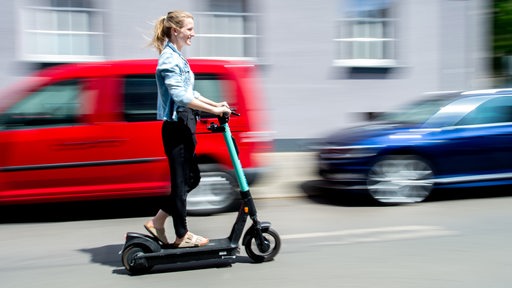 Eine junge Frau fährt mit einem türkisen Roller an parkenden Autos vorbei.