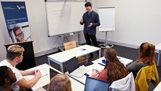 Studenten sitzen in einem Raum und hören einem Dozenten zu, der vor ihnen steht.