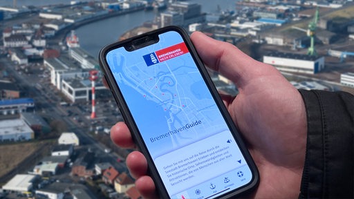 Eine Hand hält ein Smartphone mit dem Bremerhaven-Guide, dahinter der Fischereihafen (Montage)