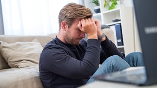 Ein junger Mann sitzt in einem Wohnzimmer und versenkt den Kopf in den Händen.