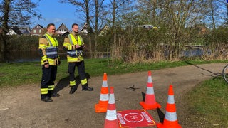 Zwei Feuerwehrmänner stehen neben einer Drohne, die in der Luft schwebt.