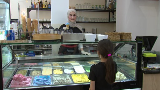 Eine Frau mit einem Kopftuch reicht einem Mädchen in einer Eisdiele ein blaues Eis