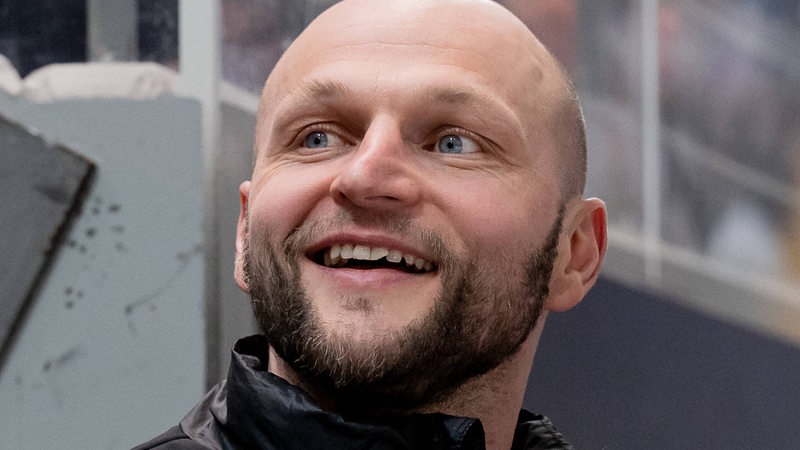 Eishockey-Manager Sebastian Furchner von den Wolfsburg Grizzlys steht strahlend an der Bande.