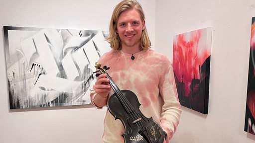 Pinguins-Spieler Moritz Wirth steht lächelnd in einer Kunstgalerie mit einer signierten schwarzen Geige in der Hand.