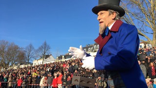 Ein Mann in Frack und mit Zylinder steht am Weserdeich. Im Hintergrund schauen ihm Hunderte Menschen auf dem Deich zu.