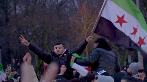 In einer Menschenmenge hält ein junger Mann eine Syrien Flagge in der Hand und zeigt das Peace Zeichen