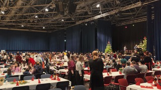 In einer Halle sitzen zahlreiche Personen an Tischen.