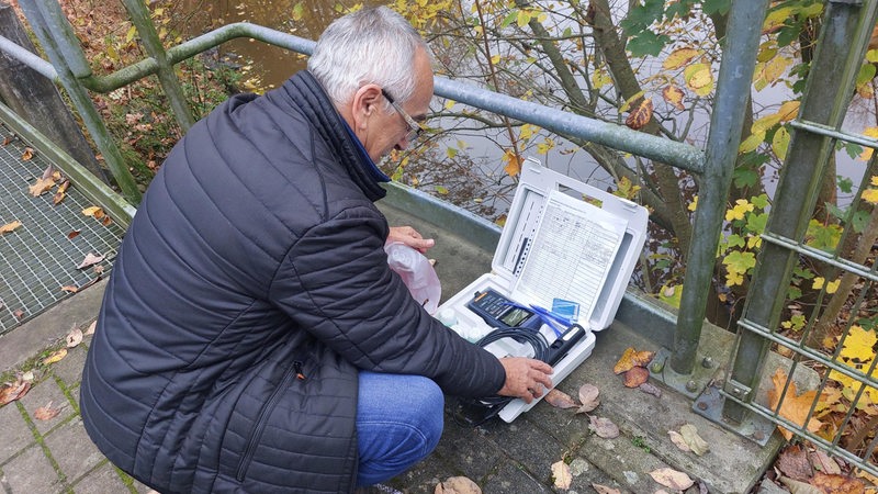 Ein Mann hockt mit einem Koffer vor einem Gewässer.