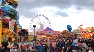 Das Riesenrad ist im Hintergrund zu sehen. Im Vordergrund laufen Besucher über den Freimarkt.