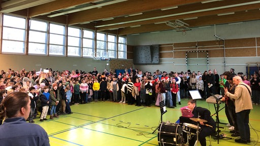 Zahlreiche Kinder und Jugendliche stehen vor einer Band in einer Sporthalle.