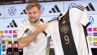 Niclas Füllkrug hält auf der Presekonfernez sein Trikot in die Kamera.