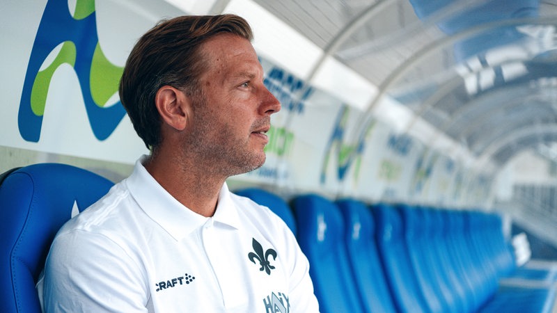 Werders ehemaliger Fußball-Trainer Florian Kohfeldt sitzt nach seiner Vorstellung bei Darmstadt 98 auf der Trainerbank und schaut nachdenklich aufs Spielfeld.