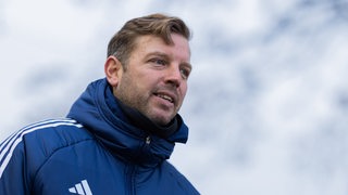 Der ehemalige Werder-Trainer Florian Kohfeldt steht in einer blauen Jacke auf dem Trainingsplatz bei seinem neuen Verein KAS Eupen.