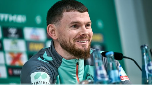 Werder-Spieler Oliver Burke sitzt lächelnd bei einer Pressekonferenz am Mikrofon.