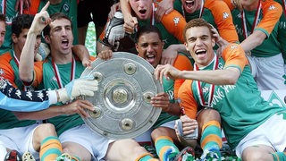 Die Werder-Spieler Johan Micoud, Ailton und Valerien Ismael sitzen nach der Saison 2004 jubelnd mit der Meisterschale auf dem Rasen.