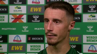 Werder-Kapitän Marco Friedl nach dem Spiel beim Interview vor einer Werbewand.