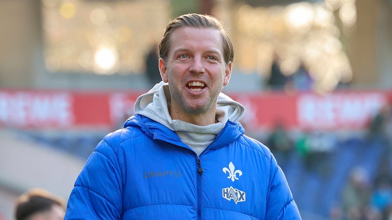 Florian Kohfeldt steht in einer Jacke des SV Darmstadt 98 an der Seitenlinie und lacht.