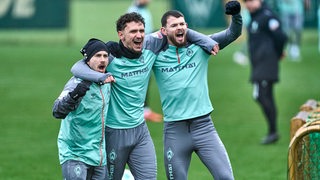 Die Werder-Profis Romano Schmid, Milos Veljkovic und Oliver Burke stehen beim Training Arm in Arm jubelnd zusammen.