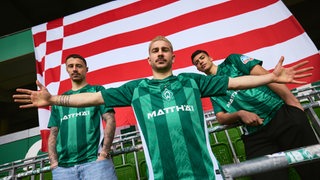 Werders Romano Schmid steht vor der Speckflagge im Weserstadion. Er trägt das neue Werder-Trikot. Hinter Schmid stehen Marco Friedl und Julian Malatina im gleichen Dress.